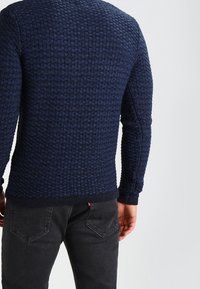 Marineblauer Strickpullover mit strukturiertem Waffelmuster, Rundhalsausschnitt und gerippten Bündchen, kombiniert mit dunklen Jeans.