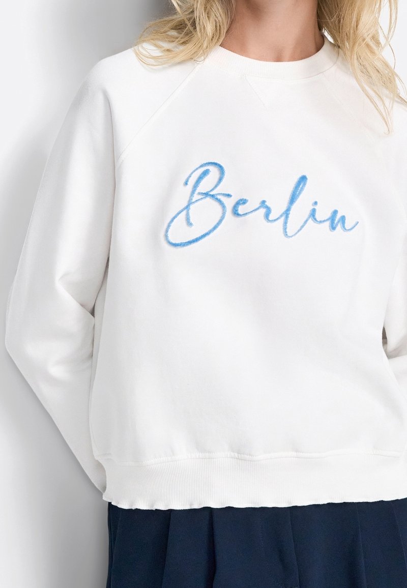 Felpa bianca con ricamo blu in corsivo della parola "Berlin". Il design presenta un colletto rotondo e polsini e orlo a costine.