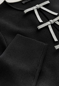 Schwarzer Strickpullover mit kristallverzierten Schleifen am Halsausschnitt und strukturierten Ärmeln. Glatter Stoff mit gerippten Kanten.