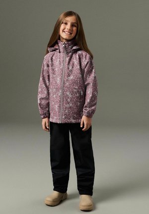 Mosolygó lány rózsaszín virágos cipzáras dzsekiben, fekete nadrágban és bézs utcai cipőben, szürke egyszerű háttér előtt áll.