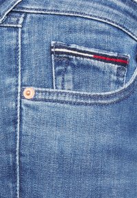 Jeans de mezclilla azul con un pequeño bolsillo trasero. La costura decorativa incluye rayas blancas, azules y rojas. Presenta un remache de cobre cerca del bolsillo.
