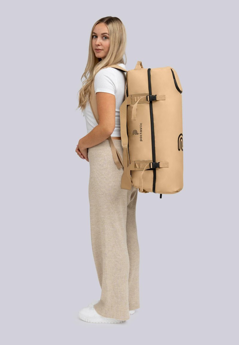 Sac à dos imperméable beige avec des bretelles ajustables, en forme arrondie, et accents noirs, comportant un logo sur le côté et une fermeture à boucle sécurisée.