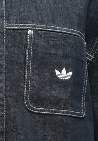 Σκούρο denim ύφασμα με ραμμένη τσέπη που δείχνει το λευκό λογότυπο trefoil της Adidas και λεπτομέρεια από ασημένιο καρφί στο επάνω αριστερό μέρος.