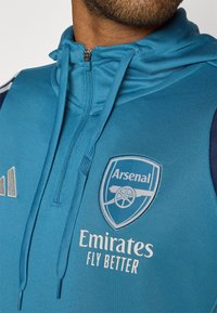 Blue sports hoodie made of a smooth material, featuring a zippered collar, drawstrings, and the Arsenal logo with white text below.