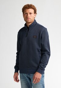 Jachetă din fleece cu fermoar, de culoare bleumarin, cu un material texturat, guler în picioare și buzunare laterale. Prezintă un mic patch cu logo pe piept.