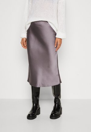 Femme portant un pull en maille blanc, une jupe midi grise brillante et des bottes hautes à talons noires, debout sur un fond clair uni.