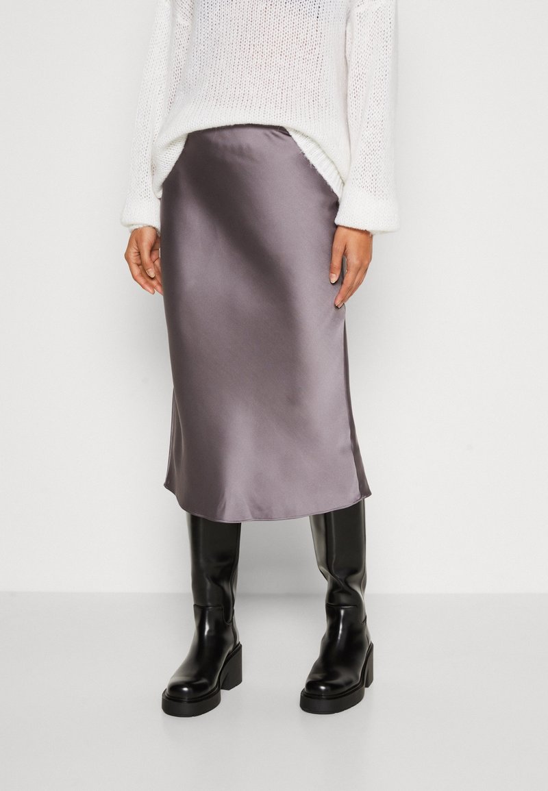 Femme portant un pull en maille blanc, une jupe midi grise brillante et des bottes hautes à talons noires, debout sur un fond clair uni.