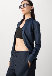 Camicia azzurra scuro con bottoni, tessuto leggero, design aderente, abbinata a un crop top nero e a pantaloni blu navy con bottoni e tasche.