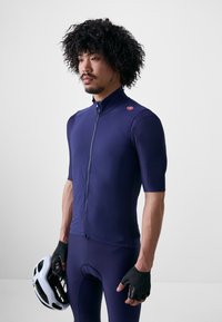 Námornícka cyklistická dres s krátkymi rukávmi, s predným zipsom, textúrovaná tkanina, priliehajúci strih, v kombinácii s čiernymi polovičnými prstami rukavicami a bielou prilbou.