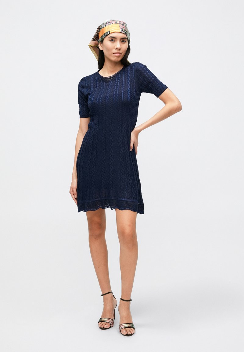 Navy knitted dress with short sleeves, textured diamond pattern, scalloped hem, paired with patterned heeled sandals and a headscarf.
