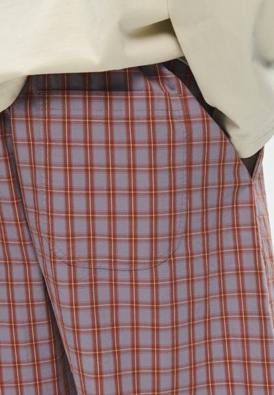 Gros plan d'une main dans la poche arrière d'un pantalon à carreaux rouge et gris, associé à un haut beige.