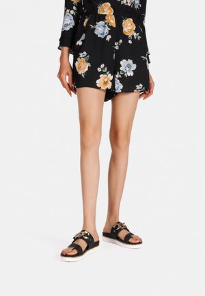 Romper nero floreale con maniche corte, caratterizzato da fiori arancioni e blu, indossato con sandali neri decorati con dettagli in metallo dorato.