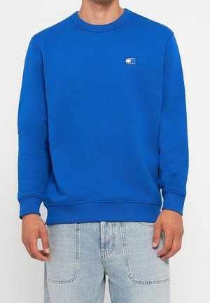 Mann trägt ein einfaches, leuchtend blaues Sweatshirt mit Rundhalsausschnitt und kleinem "Tommy Jeans"-Logo auf der Brust sowie hellblaue Jeans.