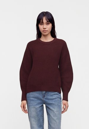 Donna con capelli neri lisci che indossa un maglione marrone scuro a maglia e jeans azzurri, in piedi davanti a uno sfondo bianco semplice.