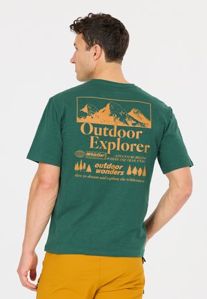 Man draagt een groen T-shirt met een oranjekleurige bergafbeelding en de tekst "Outdoor Explorer" en gele korte broek, met zijn rug naar de camera toe.