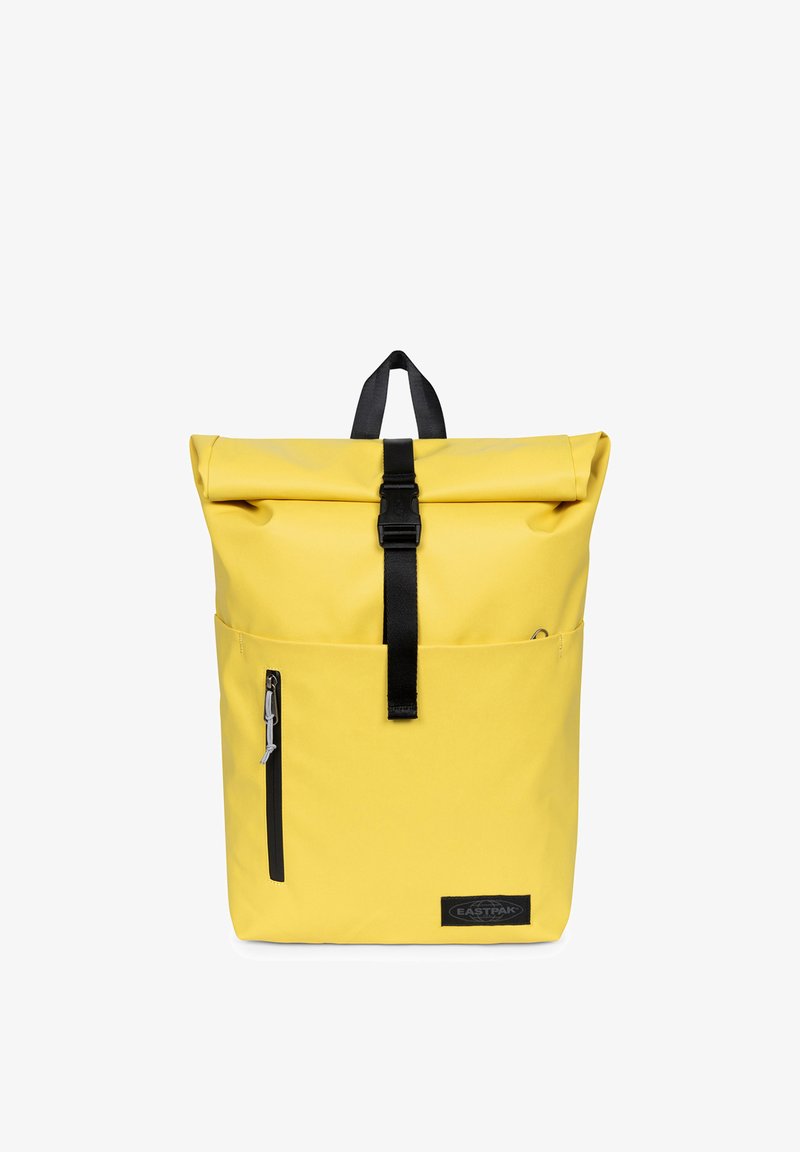Sac à dos jaune à rabat roulé avec sangle à boucle noire, poche zippée à l'avant, poignée de transport et logo Eastpak dans le coin inférieur droit avant.