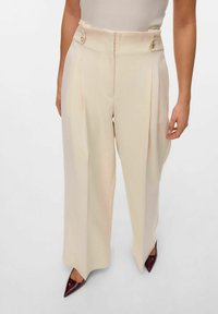 Pantalons larges en beige clair, présentant une taille haute avec fermeture à double bouton et des détails de plis. Texture lisse, tissu fluide.