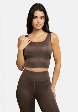 Woman with long dark hair wearing a brown sleeveless crop top and high-waisted leggings, posing with one hand near shoulder.