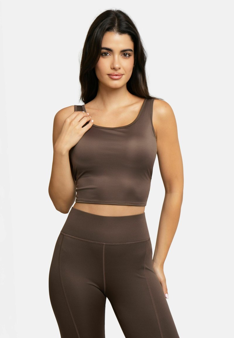Femme aux longs cheveux foncés portant un crop top marron sans manches et des leggings taille haute, posant avec une main près de l'épaule.