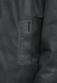 Casaco impermeável preto com textura suave e um bolso retangular com o nome "Barbour" bordado em fio escuro na parte da frente.