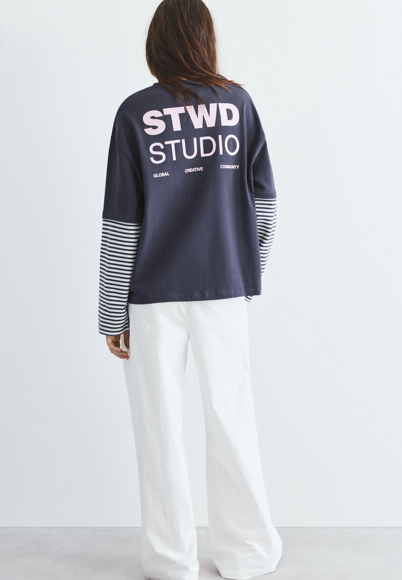 Persoană care poartă o bluză cu mânecă lungă în bleumarin cu mâneci cu dungi și textul „STWD STUDIO” pe spate, asortată cu pantaloni albi largi.