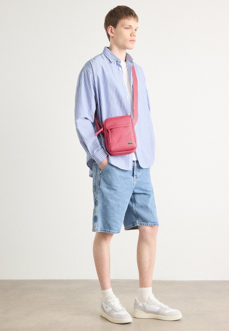 Sac en cuir rouge porté en bandoulière avec une poche zippée, associé à une chemise rayée bleue, un short en denim et des baskets de couleur claire.