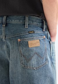 Henkilö pukeutuneena sinisiin Wrangler-farkkuihin, joissa näkyy takatasku ja logoplässi, käsi lepää etutaskussa.