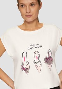 Biała koszulka z krótkim rękawem z graficznym wzorem trzech szpilek, z dodatkami takimi jak kokarda i perły, oraz napisem "tam ona ŚWIECI".