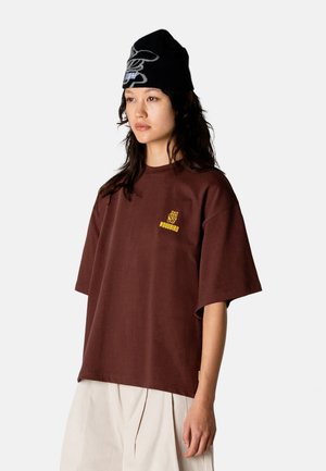 Kobieta ubrana w brązowy, oversize'owy t-shirt z żółtym logo "WOODBIRD", kremową spódnicę oraz czarną czapkę z białym wzorem na jednolitym tle.