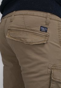 Nahaufnahme von beigen Cargohosen mit Klappentaschen und einem kleinen dunklen Etikett an der Gesäßtasche, getragen mit einem dunklen Pullover.