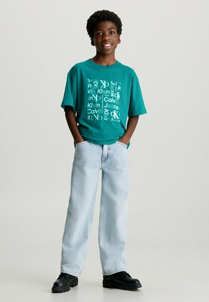Camiseta de gran tamaño color verde azulado con gráfico de texto blanco, combinada con unos vaqueros holgados azul claro y zapatos negros. El modelo está de pie con las manos en los bolsillos.