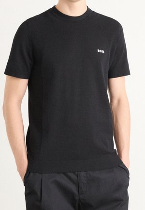 Homme portant un t-shirt noir uni à manches courtes avec le logo "BOSS" sur la poitrine gauche, mains dans les poches, sur un fond clair uni.