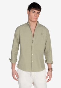 Camisa de botones verde claro con mangas largas, puños enrollables y una textura sutil. Presenta un pequeño diseño bordado en el pecho.
