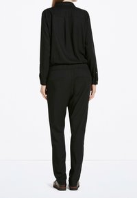 Zwarte jumpsuit met lange mouwen en een ontspannen pasvorm, elastische tailleband en een rugplooidetail. Voorzien van knoopsluiting aan de mouwen en zijzakken.