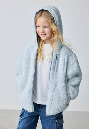 Smiling girl with long blonde hair wearing a light blue fluffy hooded jacket and blue cargo pants, standing with hands in pockets.