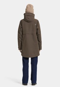 Brown waterproof parka with a cinched waist, a hood, and a rear vent. Paired with dark denim jeans and a tan ribbed beanie.