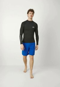 Melnas garās piedurknes rash guard ar logo uz krūtīm, apvienots ar spilgti ziliem peldkostīmiem, nēsāts basām kājām uz neitrāla fona.