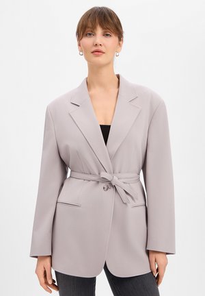 Vrouw die een lichtgrijze blazer met ceintuur draagt over een zwarte top en donkere broek, staand tegen een effen witte achtergrond.