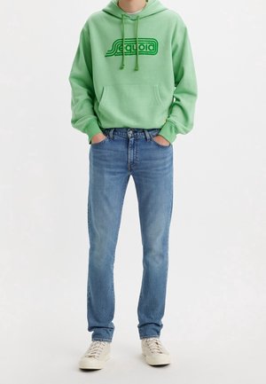 Personne portant un sweat à capuche vert clair avec le texte "Sequoia", un jean bleu et des baskets beiges, debout les mains dans les poches.