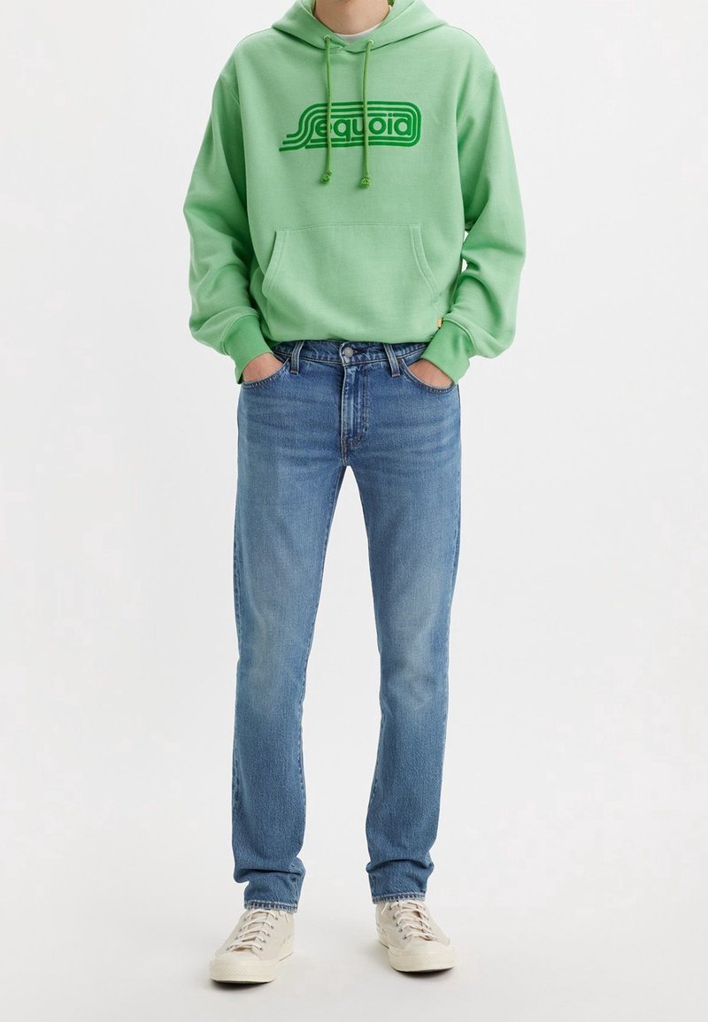 Personne portant un sweat à capuche vert clair avec le texte "Sequoia", un jean bleu et des baskets beiges, debout les mains dans les poches.
