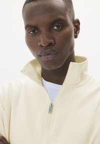 Halve-rits pullover in lichtcrème met een gladde textuur, met een zilveren rits en doorstikte naden, gelaagd over een wit shirt.
