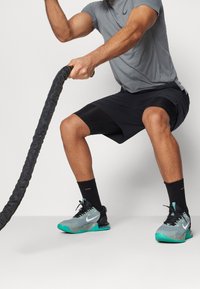 Harmaa lyhythihainen paita, mustat shortsit teksturoiduilla yksityiskohdilla, mustat sukat ja vihreät ja harmaat urheilukengät. Henkilö harjoittelee taistonköydellä.