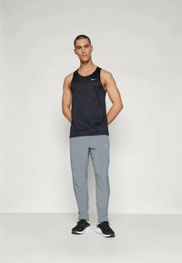 Top sport negru fără mâneci cu un logo mic, asociat cu pantaloni sport gri deschis și adidași negri. Modelul stă cu fața spre cameră, pe un fundal neutru.