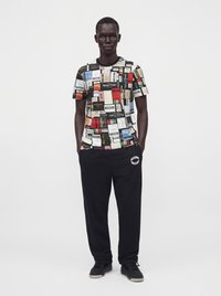Άνδρας που φοράει πολύχρωμο T-shirt Moschino patchwork με λογότυπο, μαύρη φόρμα Moschino και μαύρα αθλητικά παπούτσια, στέκεται με τα χέρια στις τσέπες.