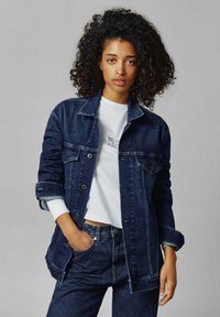Giacca di denim blu scuro, con chiusura a bottoni, tasche sul petto e maniche arrotolate. Abbinata a una maglietta bianca a collo tondo e jeans scuri.