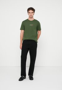 Πράσινο βαμβακερό T-shirt με εκτύπωση ορθογώνιου λογότυπου σε κόκκινο και λευκό χρώμα στο στήθος, συνδυασμένο με μαύρες παντελόνες και μαύρα παπούτσια.