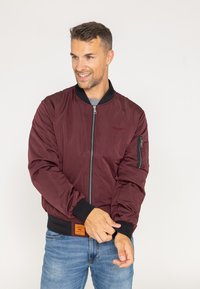 Burgundowa kurtka bomberka z czarnymi ściągaczami przy mankietach i kołnierzu. Posiada zamek z przodu oraz zapinaną na zamek kieszeń na rękawie. Gładki, lekki materiał.