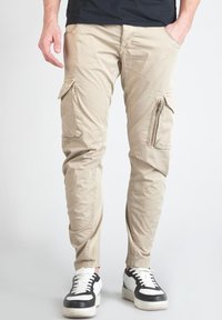 Beige cargobroek met ritszakken, tapered fit en elastische enkelmanchetten, gecombineerd met zwart-witte sneakers.
