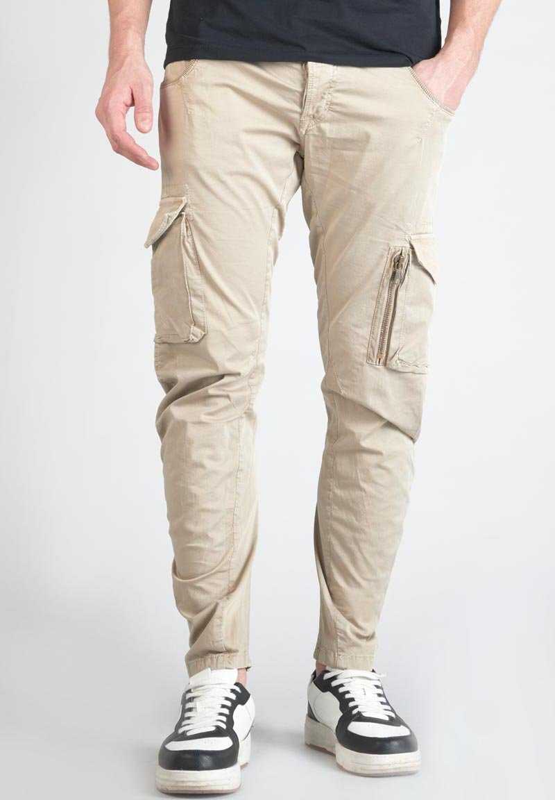 Beige cargobroek met ritszakken, tapered fit en elastische enkelmanchetten, gecombineerd met zwart-witte sneakers.