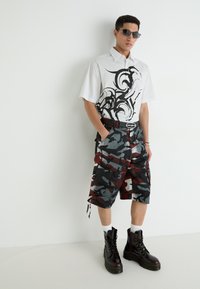 Camisa de manga corta blanca a rayas con diseño gráfico negro, combinada con pantalones cortos cargo de camuflaje multicolor y botas negras con cordones.